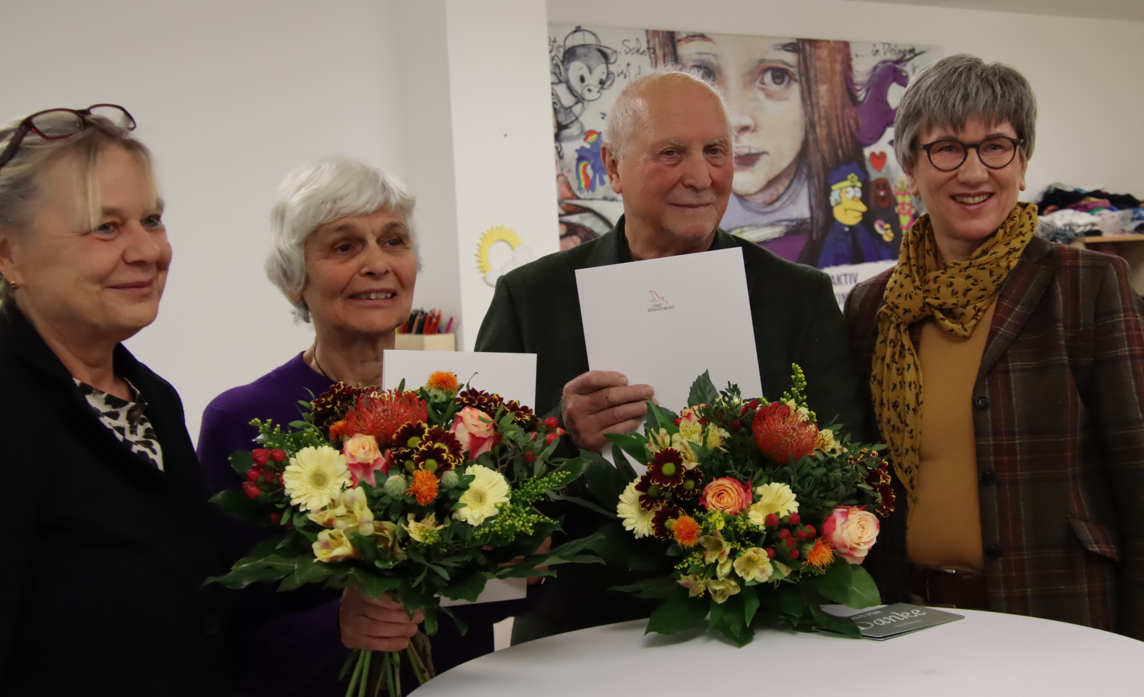 4 Personen stehen um einen Stehtisch. Ältere Frau und älterer Mann sind in der Mitte und halten Freude strahlend ihre Blumen in der Hand. Festlicher Anlass.