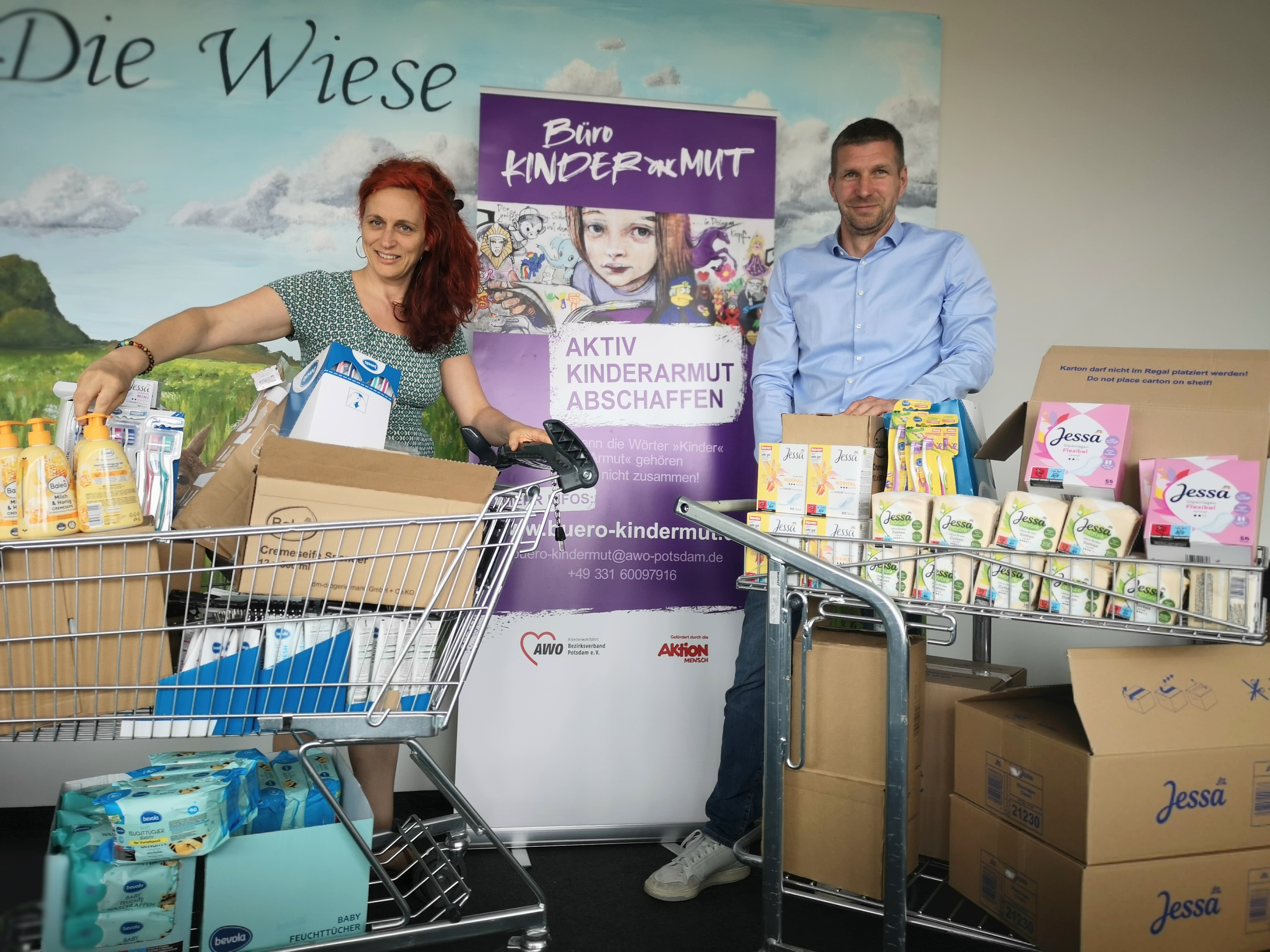 Im Büro Kinderarmut, eine Frau und ein Mann, jeweils mit einem vollen Einkaufswagen mit Hygieneartikeln zwischen ihnen das lila Roll-Up vom Büro Kinderarmut.