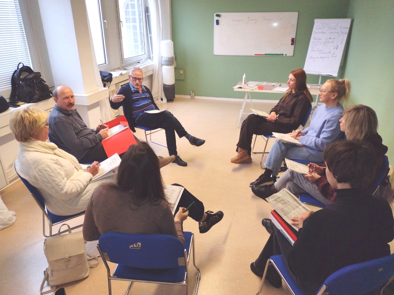 8 Menschen sitzen im Kreis sitzen. Alle haben etwas zum Schreiben auf dem Schoß, eine Tafel ist an der hinteren Wand.