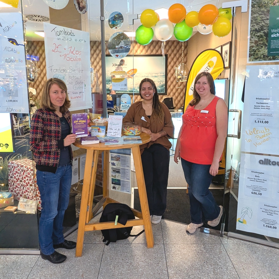 Vor einem Reisebüro. Ein Stehtisch an dem drei Frauen stehen und Lose verkaufen.