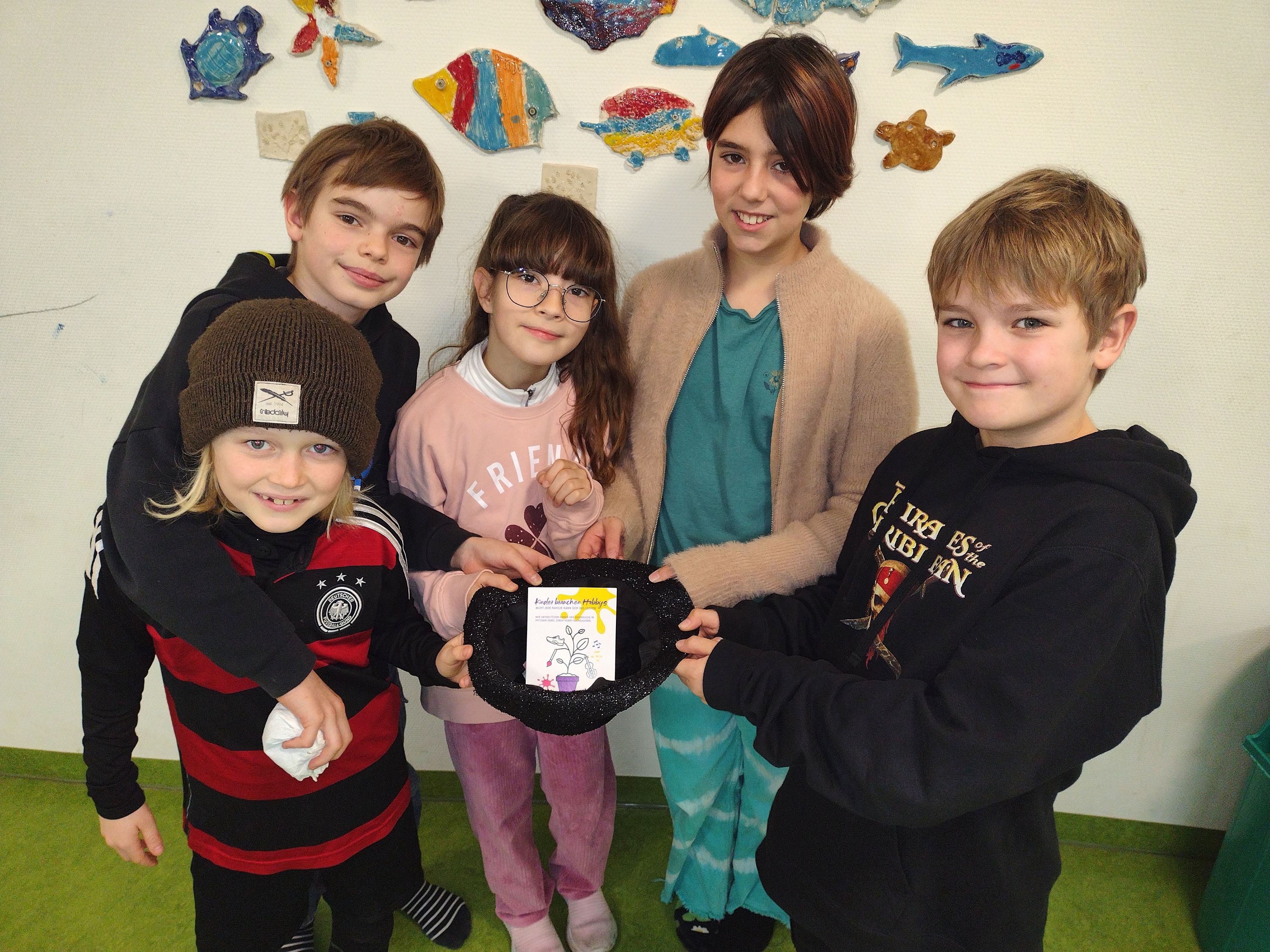 5 Kinder eng beieinander halten eine Mütze in die Mitte, darin eine Karte. Die Kinder sind fröhlich.