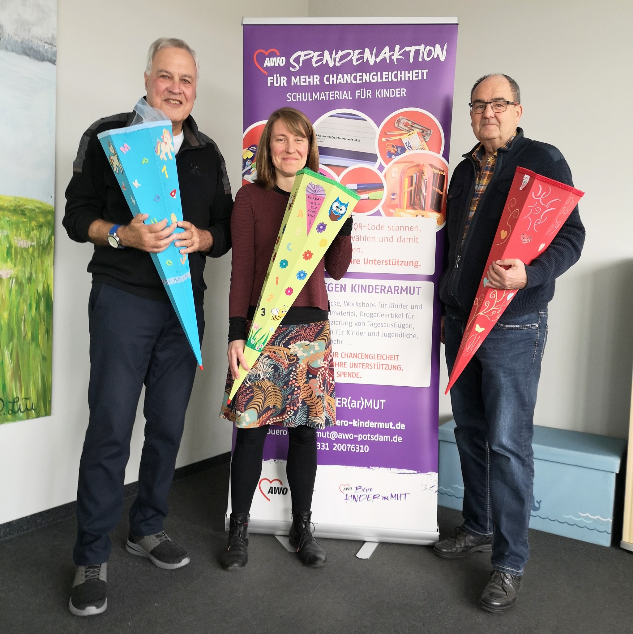 Zwei Männer und eine Frau halten große Zuckertüten in den Händen und lächeln. Sie stehen vor einen lila Aufstteller vom Büro Kindermut.