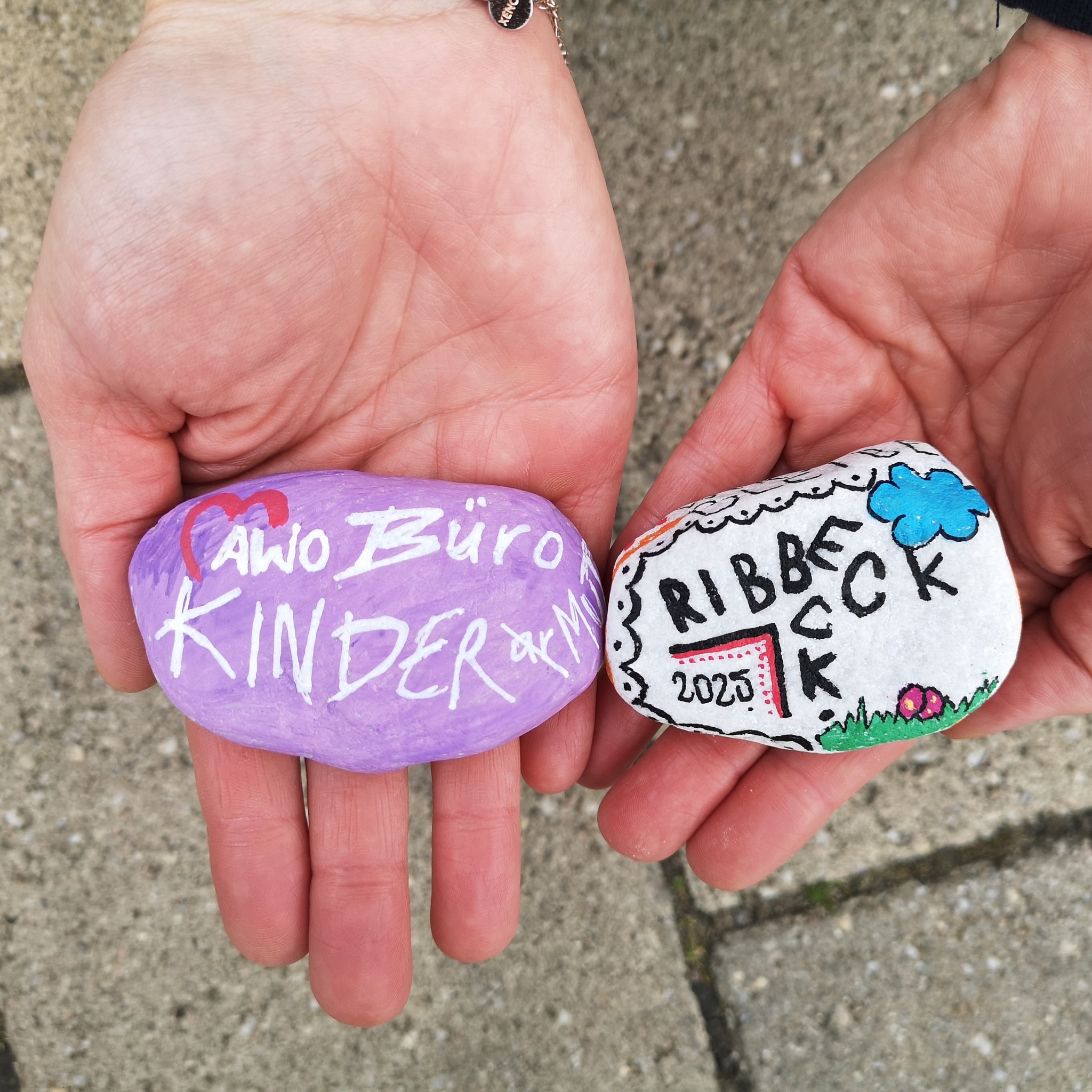 Zwei Hände auf denen jeweils ein bemalter Stein liegt. links: lila Stein mit weißer Schrift: AWO Büro Kindermut   rechts: weißer Stein mit schwarzer Schrift: Ribbeck 2025 mit Wolke und Wiese.