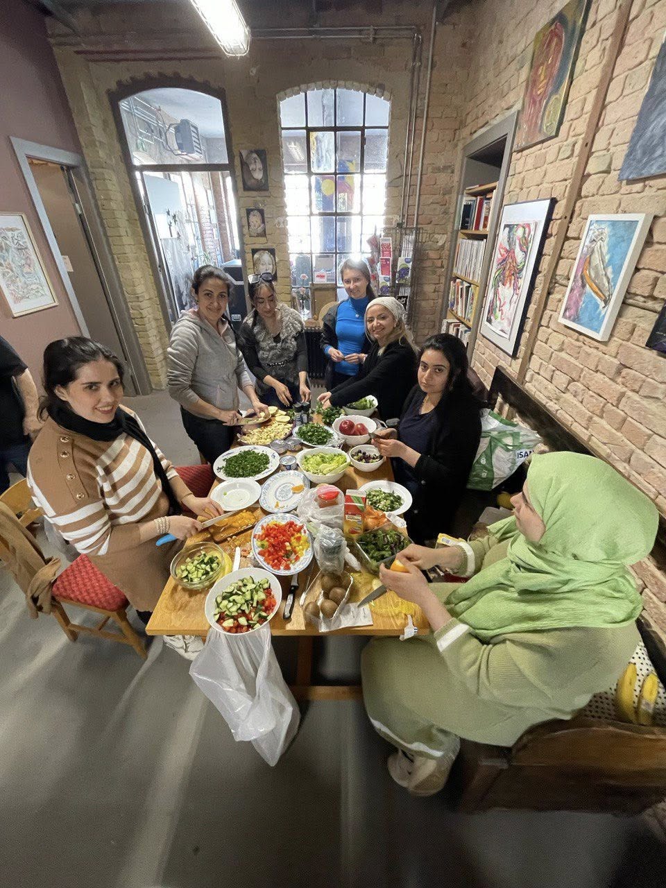 Raum mit Steinmauern und Bildern an der Wand. Voller Tisch, Frauen bei der Zubereitung einer Mahlzeit. Gutgelaunte Gemeinschaft.