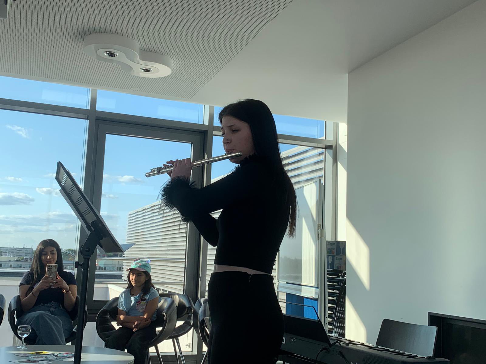 Junge Frau/Mädchen mit Flöte, vor ihr ein Notenständer. Glaswände bieten einen tollen Ausblick. Zwei weitere Mädchen sitzen auf Stühlen.