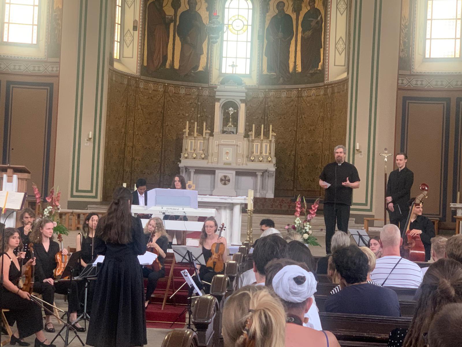 In einer Kirche. Junge Menschen mit ihren Intrumenten in festlicher schwarzer kleidung. Dirigentin von hinten mit langen dunklen Haaren.