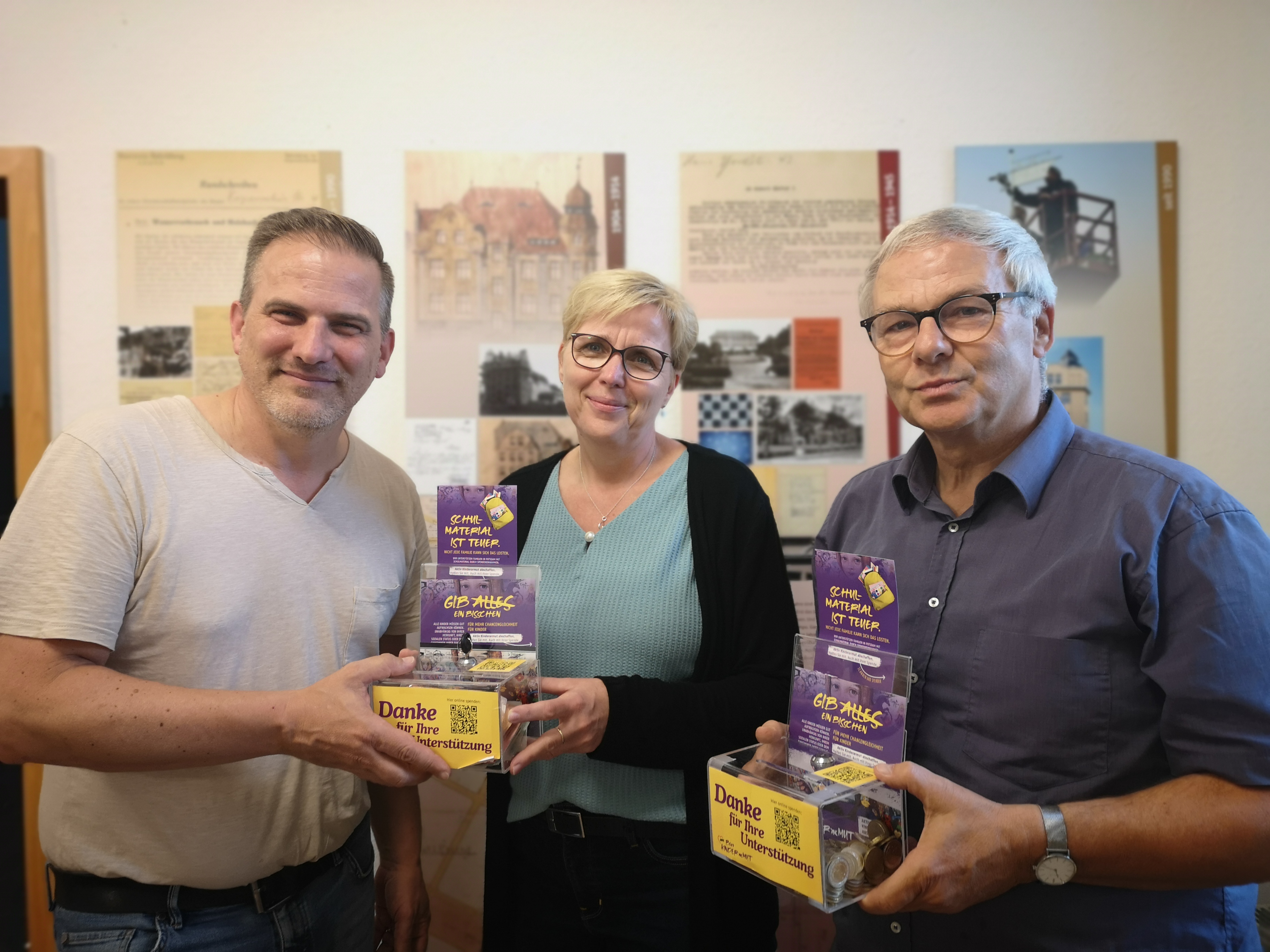 3 Menschen mit zwei Spendenboxen in den Händen. In der Mitte steht eine Frau. Alle lächeln in die Kamera.