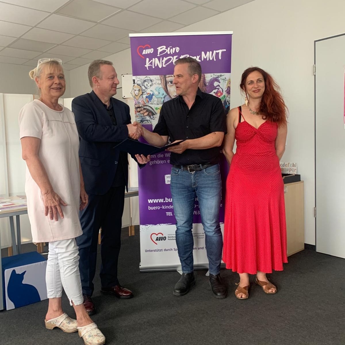 4 Personen vor einem Roll-Up des Büro Kindermut. 2 Frauen außen, innen schütteln sich die Männer die Hand.