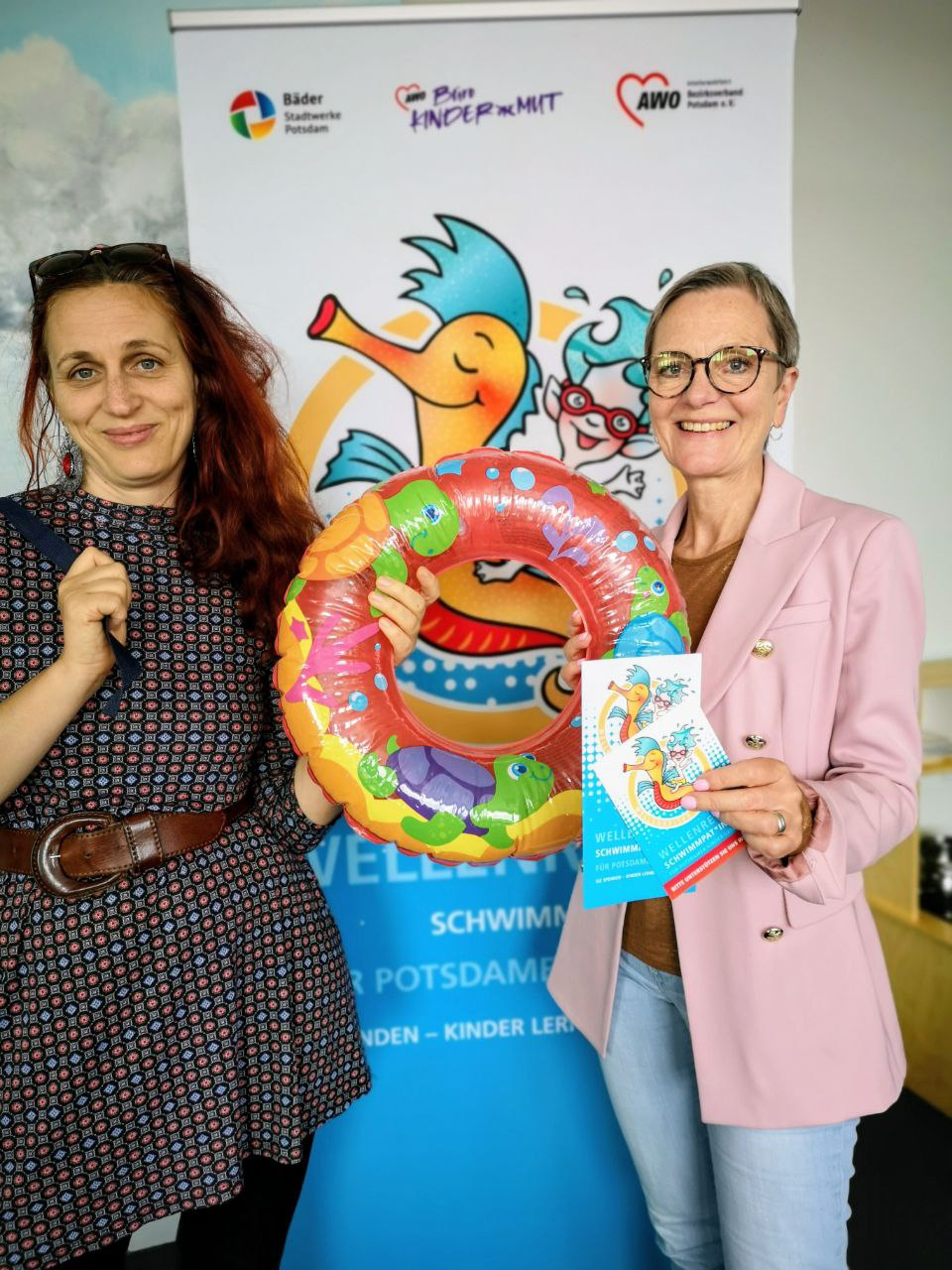Zwei Frauen mit einem bunten Kinder-Schwimmreifen zwischen sich stehen vor dem Aufsteller des Projektes Wellenreiter