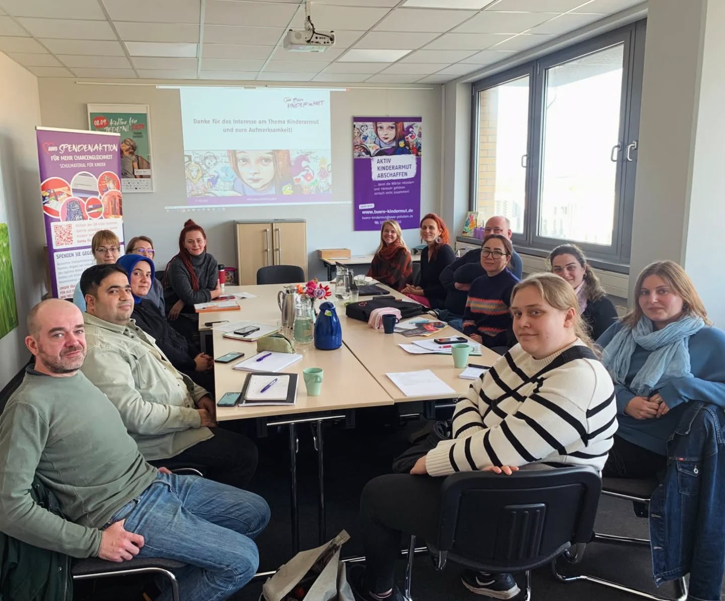 Großer Konferenzraum des Büro Kindermut. 13 Personen sitzen um einen großen Tisch und sehen in die Kamera. An der hinteren Wand läut eine Präsentation.
