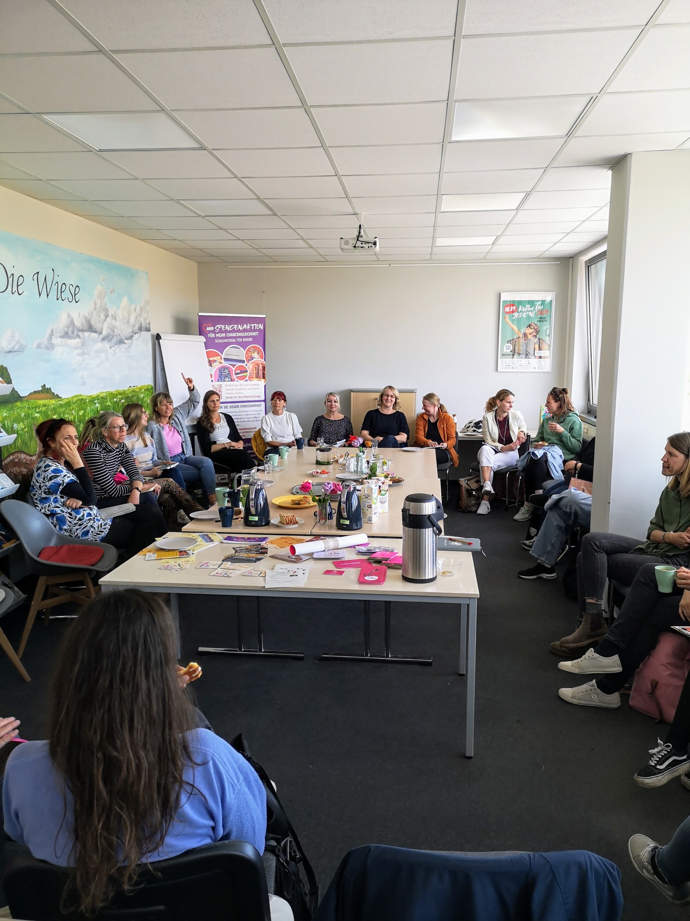 Großer heller Raum. Viele Menschen sitzen in um großen Tisch. Auf dem Tisch Getränke.